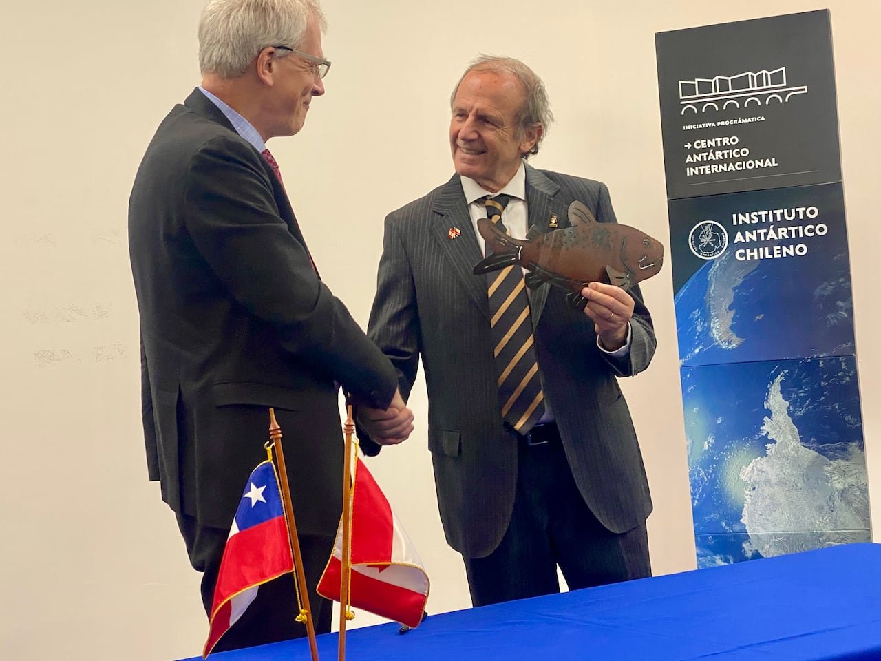 Two men in suits shake hands at a table with the Chilean and Canadian flags, celebrating an MOU between the two countries.