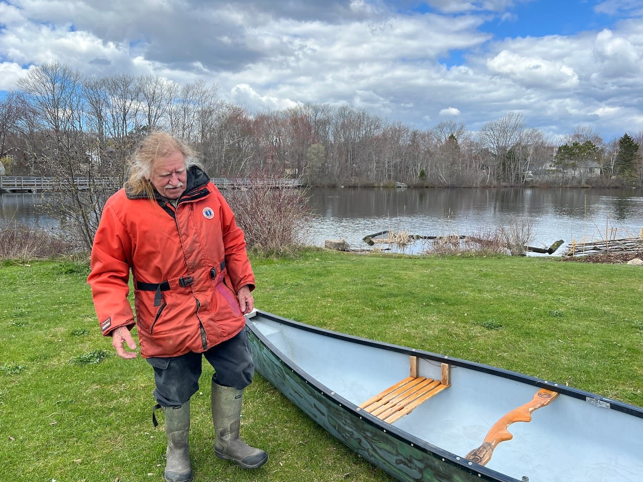 A man with white hair and an orange jacket next to a small boat in the grass.