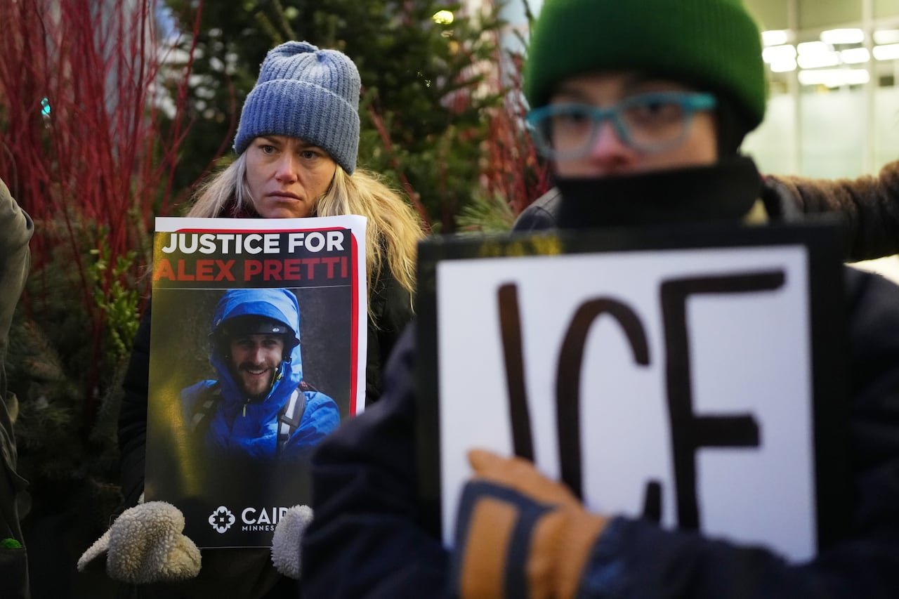 A woman with long blonde hair, wearing clothing, stands outside holding an image of a man. The sign says 'Justice for Alex Pretti."