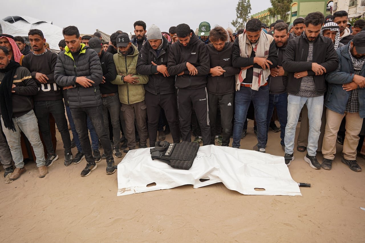 Mourners gather around a shrouded body marked 'Press' at a funeral.