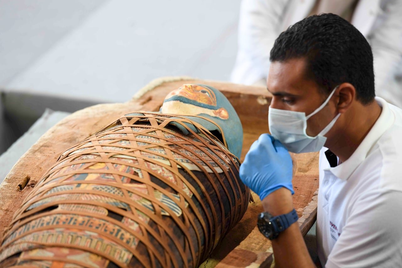 default-285 An archaeologist inspects an Egyptian mummy that is wrapped in an elaborate and colourful burial shroud. The archaeologist is wearing a medical mask and gloves.