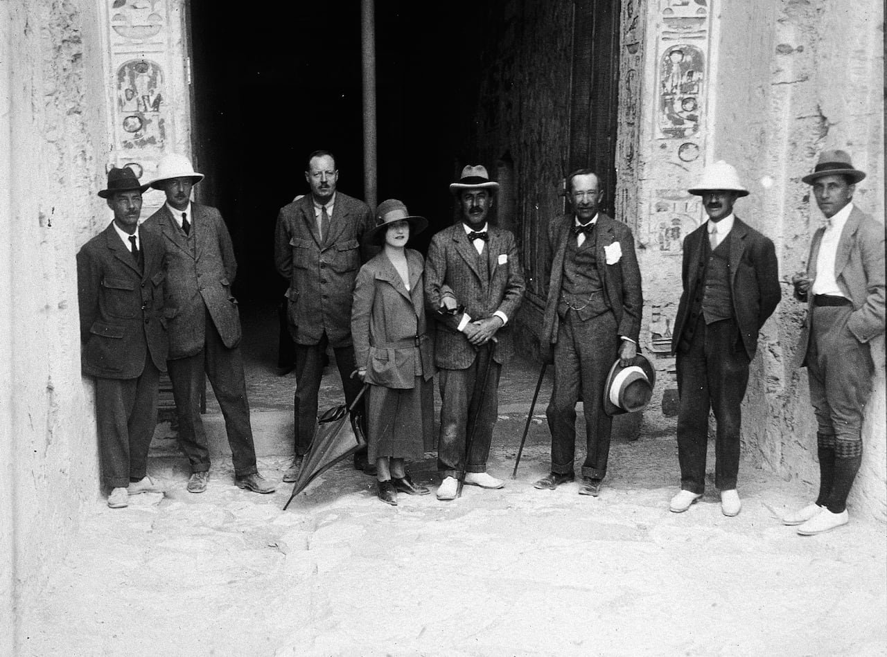 default-287 Seven men and one woman stand in front of the entrance of a stone tomb. The photograph is in black and white.