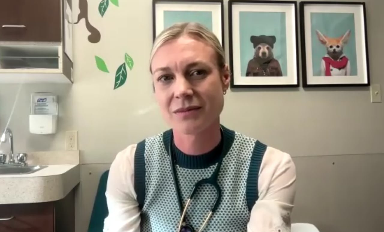 A woman with blond hair, pulled back, is wearing a black and white vest and stethoscope. A sink and animal pictures can be seen behind her.