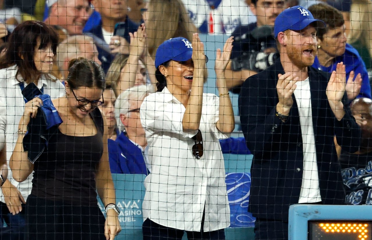 default-207 Meghan Markle in a white baseball jersey and blue ball cap stands next to Prince Harry in navy sports jacket, white t-shirt and blue ballcap as the clap and cheer in the stands of a baseball stadium