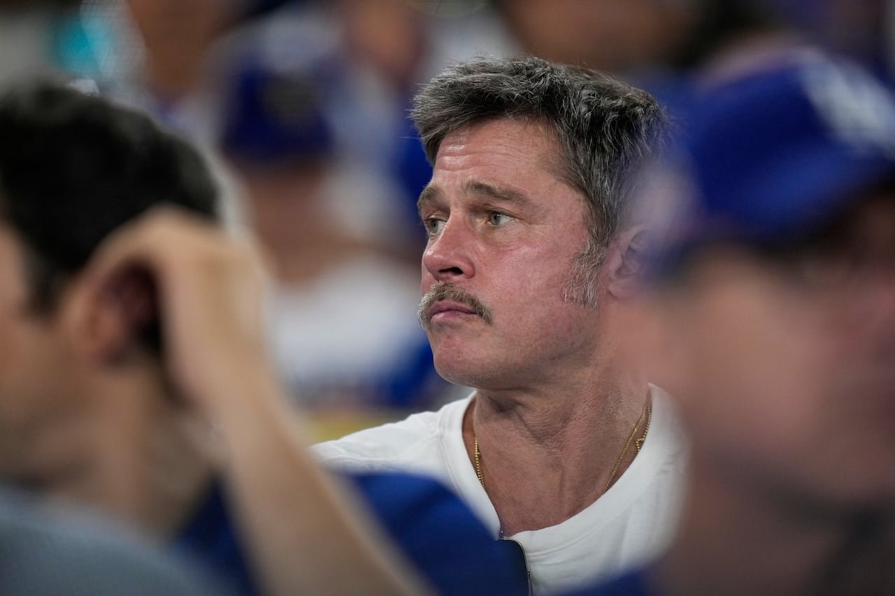 default-208 A man with a mustache, wearing a white t-shirt, is seen sitting in the stands of a stadium.