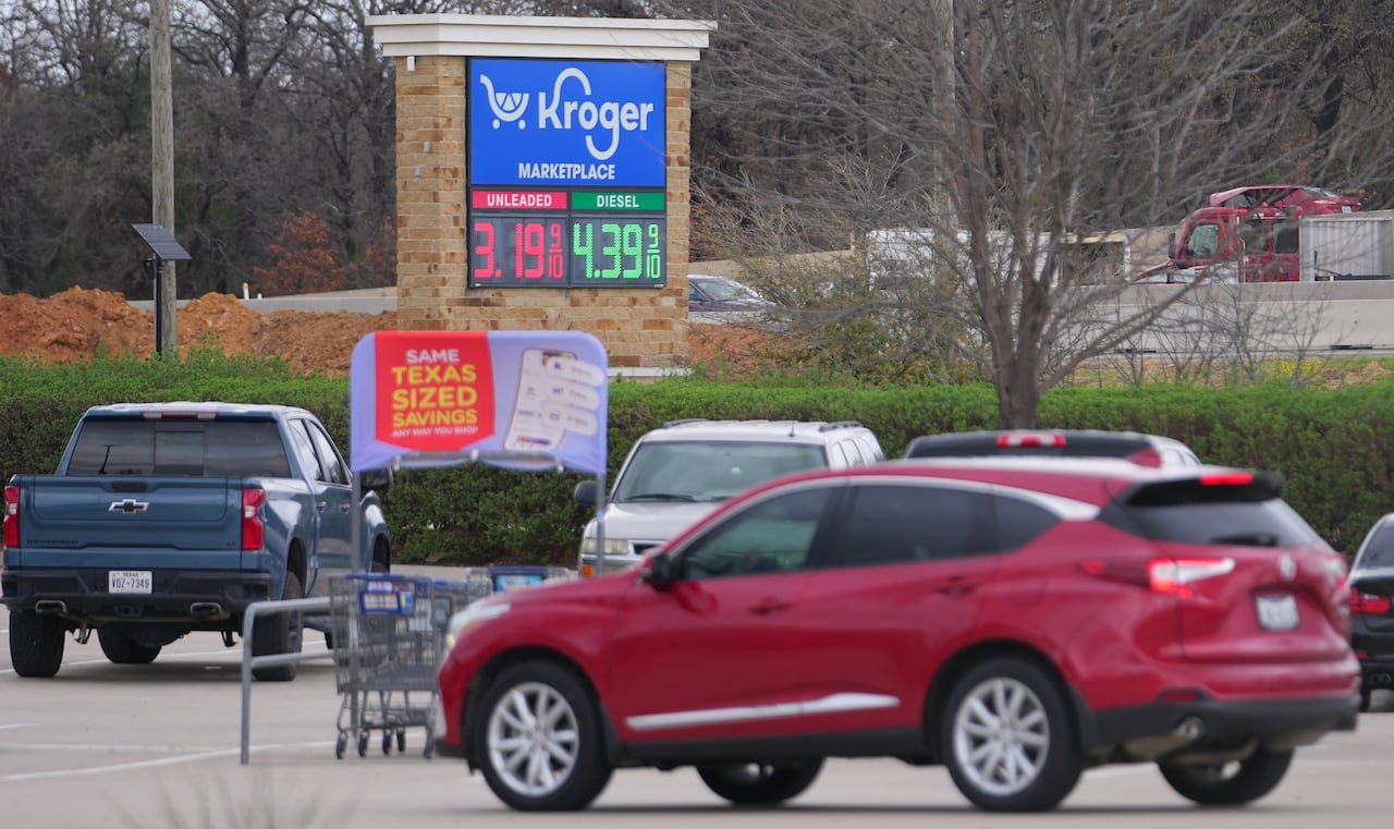 A red SUV drives through a parking lot in front of a sign that shows a price of $3.19 for a gallon of gas.